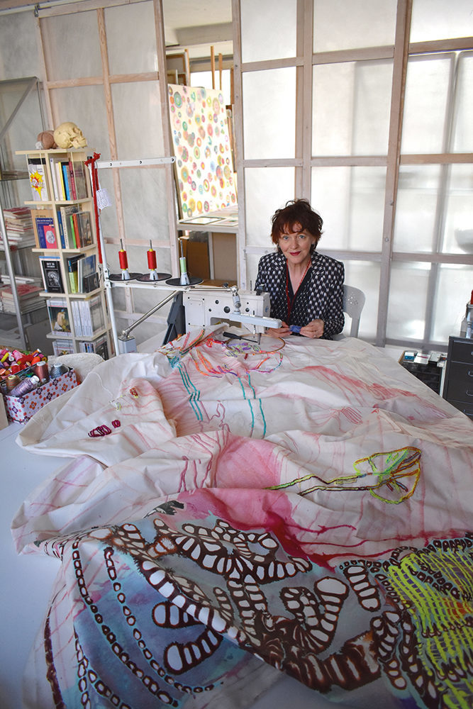 Seet van Hout im Atelier. Sie sitzt an einer Nähmaschine und arbeitet an einem bemalten Leinentuch, welches bunt bemalt ist