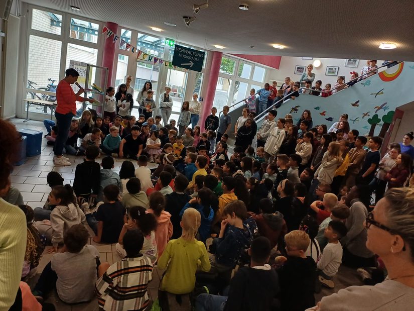 Die Schulgemeinschaft ist in der Aula versammelt. Die Zuschauer sitzen auf dem Boden. Frau Schäfers ruft mit Mikrofon die einzelnen Kinder auf.