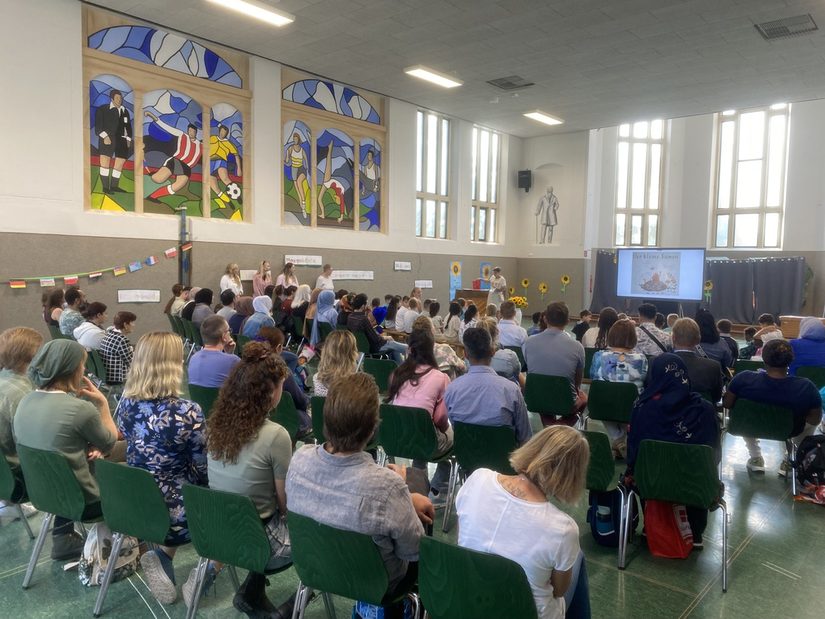 In der Aula am Theodorstandort. Frau Schäfers heißt die neuen Erstklässler willkommen. Die Eltern sitzen in Stuhlreihen und die Kinder davor.