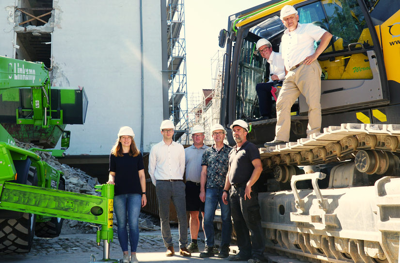Auf dem Foto sind mehrere Projektverantwortliche zu sehen, die in, auf und um einen Bagger vor der Baustelle am Abdinghof stehen.