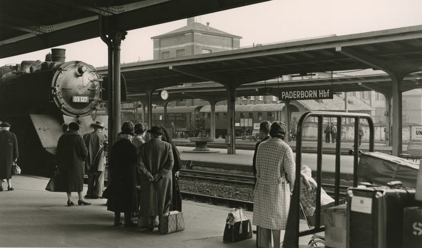 Menschen an einem Bahnhof mit einer Lokomotive und ein Schild mit der Aufschrift: "Hauptbahnhof"